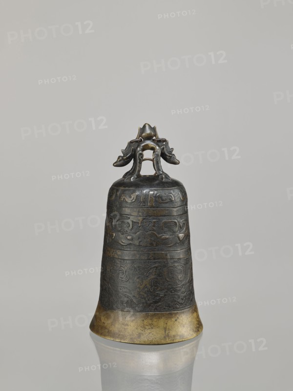 Bell, ; dragons iconography; Chinese art; Chinese culture; Qing dynasty 1644–1911; dragon with pearl; taotie iconography; meander ornament; leiwen ornament; 18th century; China place of origin; height 28.0 cm; width 15.3 cm; weight 2.29 kg, A single metal bell with a dark aged surface and a golden lower rim stands upright against a plain light gray background, featuring an ornate handle shaped like a stylized creature with curled limbs and decorative motifs; the bell body shows banded raised patterns and intricate carved designs in darker and slightly greenish-brown tones, and the base reflects faintly on the smooth surface beneath.