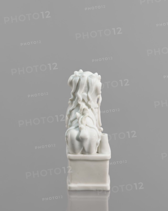 Fo lion figurine with a ball;, lion iconography; ceramics; blanc de Chine porcelain; figurine; Chinese art/culture; animal figurine; 18th century, China; sculpture; height 13.2 cm, width 4.8 cm, depth 6.7 cm., A glossy white ceramic figurine sits on a rectangular base against a neutral gray background. The figurine features flowing, wavy forms that cascade down the back, creating a sense of layered, ribbon-like texture. The base and the figure share the same smooth, reflective white surface, with subtle highlights and soft shadows emphasizing the curves and folds. The overall composition is centered in the frame, with the gray background appearing uniform and uninterrupted. Colors visible are white and gray.