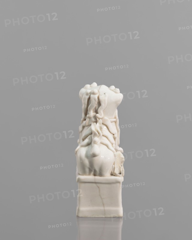 Incense burner in the shape of a fo lion with a ball; ceramic sculpture; Chinese; 18th century; height 13.0 cm, width 6.0 cm, depth 4.8 cm; censers/incense stand; animal figurine; lion iconography; fo iconography., A small glossy white sculptural object sits on a rectangular base against a plain gray background; the object has a smooth, rounded lower section and an intricately detailed upper portion with flowing, ribbonlike forms and bulbous knots that cascade downward, creating a layered, draped appearance, and there are subtle hairline cracks and slight discolorations visible in the white surface.