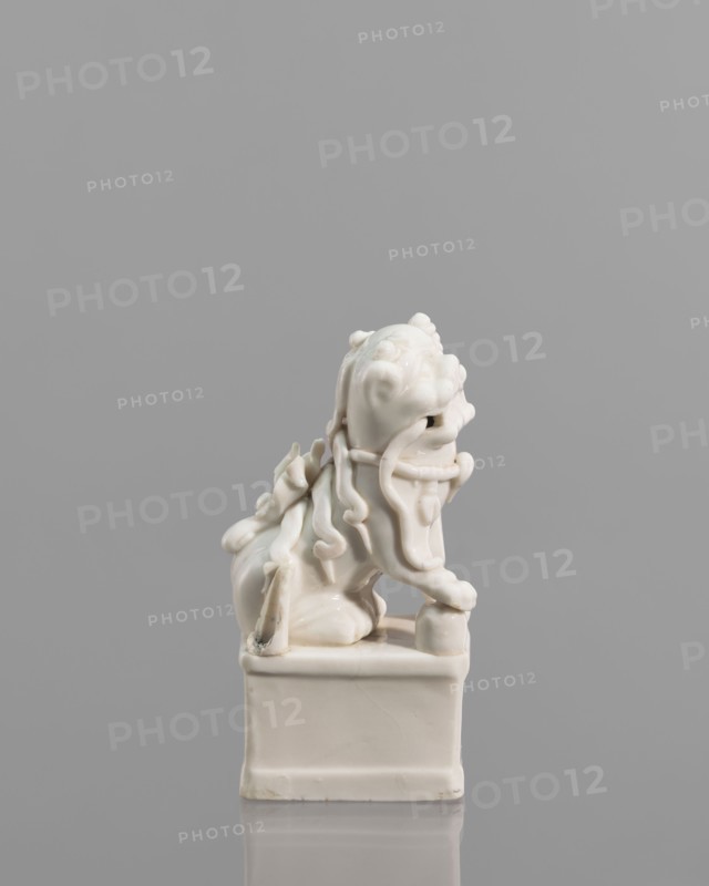 Incense burner in the shape of a fo lion with a ball; ceramic; Chinese; 18th century; sculpture; incense censer/stand; animal figurine; height 13.0 cm, width 6.0 cm, depth 4.8 cm; maker, inscription: none noted., A glossy white ceramic figurine of a stylized lion-like creature sits on a square pedestal against a smooth gray background; the creature has flowing, ribbon-like mane elements and curled details, a rounded head with pronounced brows and snout, a raised forepaw resting on a small rounded object, and a muscular body with a curled tail, all rendered in smooth, shiny white with soft shadows and faint reflections on the pedestal and surface.