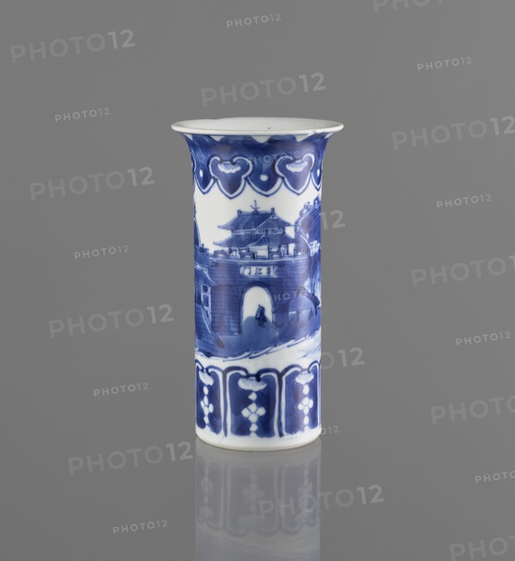 Flute-type vase, Chinese blue-and-white porcelain, landscape motifs; Kangxi period 1662–1722, 17th–18th century; China; vessel; height 15 cm, diameter 8.1 cm., A cylindrical ceramic vessel stands upright against a gray background, decorated with blue-painted scenes and patterns on a white base. The upper rim flares outward slightly and features a band of repeating heart-shaped motifs in deep blue. Below this, a painted scene shows architectural forms resembling a multi-tiered gate and surrounding structures, rendered in varying shades of blue with brushstroke textures and a small human silhouette visible near an archway. The lower section has a band of repeating vertical panels with floral or geometric motifs in dark and medium blue. The vessel casts a subtle reflection on the glossy surface beneath it. Colors visible include blue, white, and gray.
