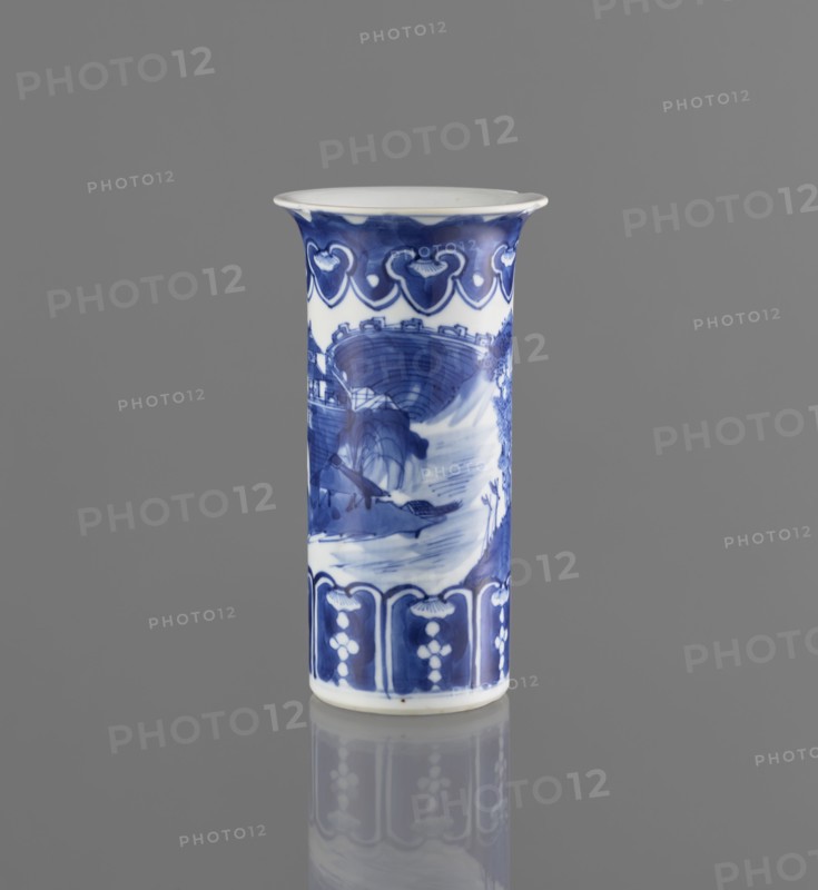 Flute-type vase, ceramics, Chinese blue-and-white porcelain with landscape motifs, Chinese, Kangxi 1662–1722, 17th–18th century, China; vessel; height 15 cm, diameter 8.1 cm., A tall cylindrical porcelain vase stands upright against a plain gray background, displaying a painted blue-and-white design; the rim flares outward slightly and is white, while a band of deep blue with lighter blue decorative motifs circles near the top; the main body shows a scene in varying shades of blue with brushwork suggesting curved architectural forms and flowing water, and a band of darker blue panels with lighter decorative elements wraps near the base; the vase sits on a reflective surface that mirrors its blue-and-white colors.