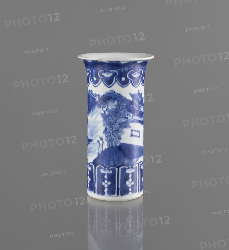 Flute-type vase, blue and white porcelain, landscape motifs, Chinese, Kangxi 1662–1722, 17th/18th century, China; vessel; height 15 cm, diameter 8.1 cm., A tall cylindrical ceramic vase stands upright against a smooth gray background, decorated in blue and white. The rim flares outward slightly and is white on top with a band of deep blue beneath, featuring a repeating scalloped motif. The central body shows a painted scene in blue tones with a landscape including a tree, hills, and architectural forms. The lower section has a decorative border in dark blue with vertical panels and small floral-like elements. The vase sits on a reflective surface that mirrors the blue and white shapes and the gray tones of the surroundings.