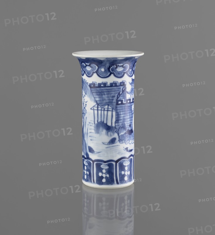 Flute-type vase, ceramics, blue-and-white porcelain, Chinese art, landscape motifs, Chinese, 17th/18th century, China, vessel, height 15 cm, diameter 8.1 cm, A tall cylindrical ceramic vase stands upright against a solid gray background, painted in white with blue decorative motifs; the rim flares slightly outward and is white on top with a blue border beneath, a band of stylized cloud-like blue patterns encircles the upper section, while the main body features a blue landscape scene with architectural shapes, sloped rooftops, leafy elements, and rounded forms, and the lower section shows a repeating blue panel design with floral-like white shapes; the vase sits on a reflective surface that shows a muted mirror image of its base and lower decorations.