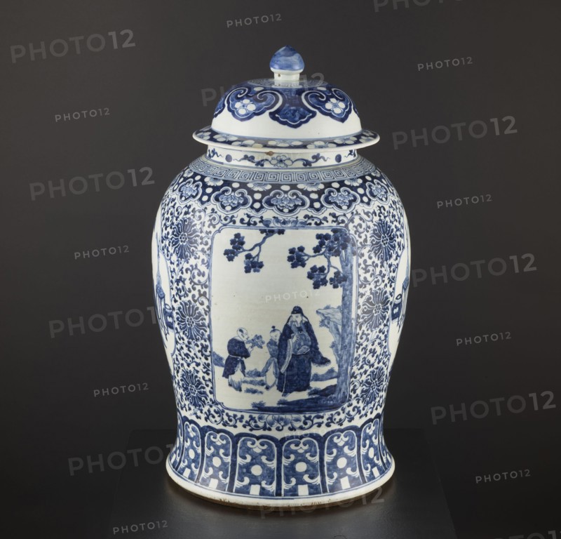 Pot with cover, resource pots, blue-and-white porcelain, Chinese art, plant motifs and mountain landscapes, decorative vessel, ceramics, Qing style, Qing 1644–1911 and Ming 1368–1644 associations, possibly 1st half of the 17th century, China, height 60 cm, diameter 36 cm., A lidded ceramic jar with a rounded body and domed lid sits against a dark background; the jar is predominantly white with extensive blue painted decoration, including floral and cloud-like motifs around the shoulder and a framed rectangular scene on the front showing human figures and tree branches; the lower body features a band of repeating scroll and dot patterns and the lid has complementary blue ornamental designs and a blue-tipped finial.