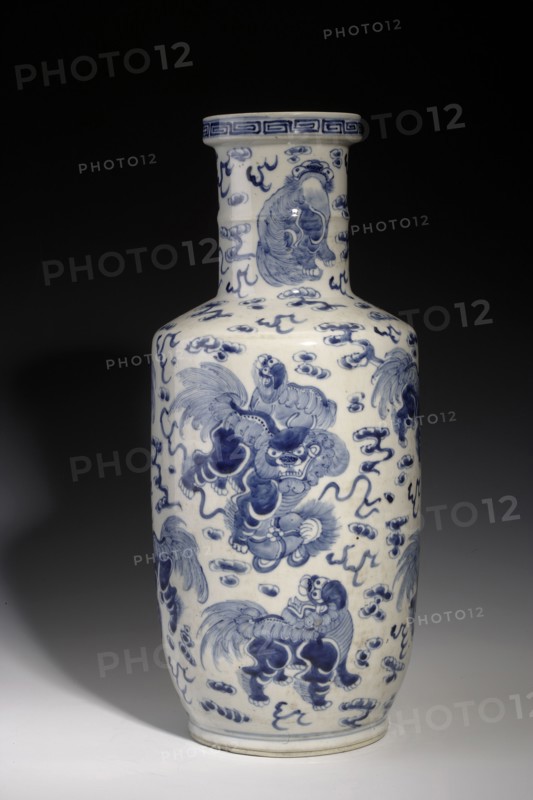 Tall bang chui vase, Chinese, Qing dynasty, Kangxi period 1662–1722, 17th century. Blue-and-white porcelain vessel shaped like a wooden hammer/pestle with a wide cylindrical neck terminating in a round ring, angular shoulders and an oval belly tapering toward the base; height 46.0 cm. Surface densely painted with fo lion motifs playing with a rag ball among clouds; ring around the neck decorated with a meander., A tall ceramic vase with a cylindrical neck and rounded body sits against a dark background, decorated in blue on a white ground with a repeating motif of stylized mythical creatures and swirling cloud-like forms; the blue decoration varies in tone from deep cobalt to lighter blue washes, and the white ceramic shows a glossy surface with subtle reflections.