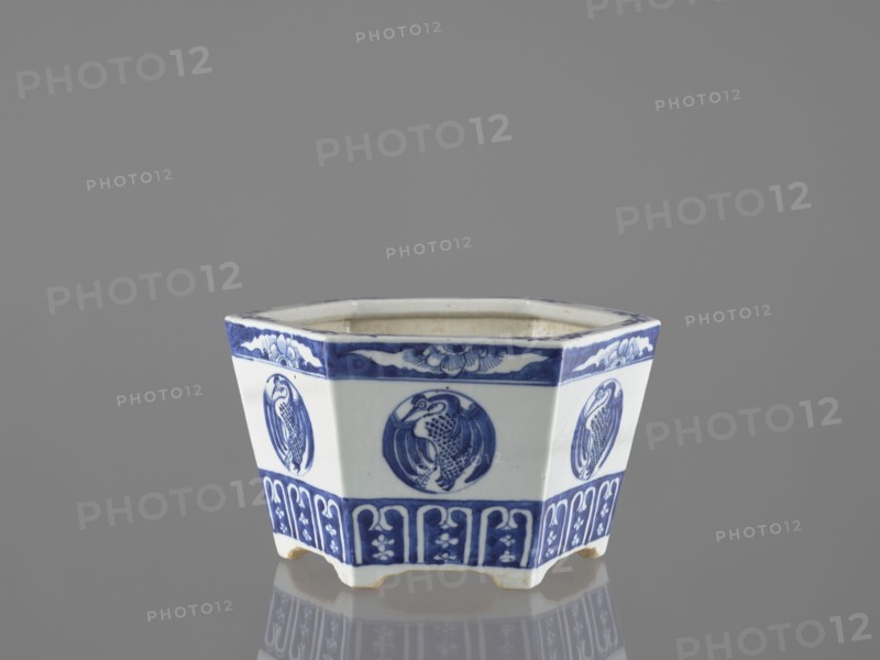 Flowerpot, Chinese, Qing dynasty 1644–1911, 18th–19th century. Blue-and-white porcelain jardinière decorated with floral motifs—lotuses and other flowers—and birds including phoenixes. Vessel, height 15.2 cm., A hexagonal ceramic container with a glossy white base and blue painted decoration sits on a reflective surface against a gray background; each side features a circular blue medallion with a bird motif and a patterned blue band near the top and bottom, and the piece rests on short feet with visible wear at the edges.