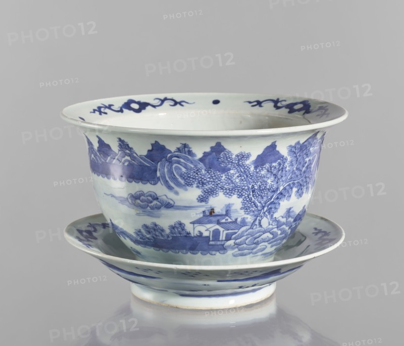 Flowerpot on base, Chinese, Qing dynasty 1644–1911, blue-and-white porcelain jardinière with landscape motifs and floral decorations, flowers, 18th–19th century, China; vessel, height 19.7 cm, diameter 28.2 cm., A ceramic bowl with a matching saucer, both decorated in blue on a white ground; the bowl has a flared rim and a rounded body sitting on a short foot, and the saucer mirrors the bowl's curved profile. The painted decoration shows a landscape scene wrapping around the bowl with a group of trees, a small building near the water, stylized cloud and wave motifs, and distant mountain shapes, while the rim and saucer edge are accented with ornamental scroll-like motifs. Visible colors include shades of blue and white, with a small area of brownish discoloration.