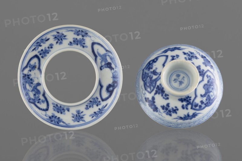 Bowl on a base with a lid;, Blue-and-white porcelain vessel, 19th century, South China, China. Figural decorations depicting female images. Height 8.6 cm., A pair of circular ceramic objects with white bases and blue decorative floral and ornamental patterns are displayed on a gray background, one shown flat with a central open ring and the other shown upside down revealing a recessed base; the blue designs include stylized flowers, leafy sprigs, and ornate cartouche-like shapes, with a narrow blue band near the inner and outer rims and a faint reflection of the objects visible on the surface below, featuring colors of white, blue, gray, and a subtle hint of off-white on the ceramic edges.