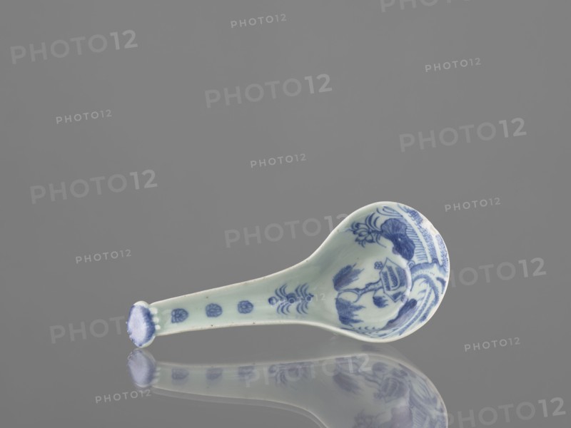 Sauce boat spoon, Chinese, blue-and-white porcelain with plant ornaments, tableware element, ceramic vessel, Qing dynasty mid-19th century, China; length 19.2 cm., A small ceramic spoon lies on a reflective surface, showing a mirrored reflection beneath it. The spoon is pale with blue decorative patterns concentrated on the bowl and along the handle, including floral and abstract motifs. The background is a uniform gray, and the reflection shows the same pale and blue colors reversed on the surface.