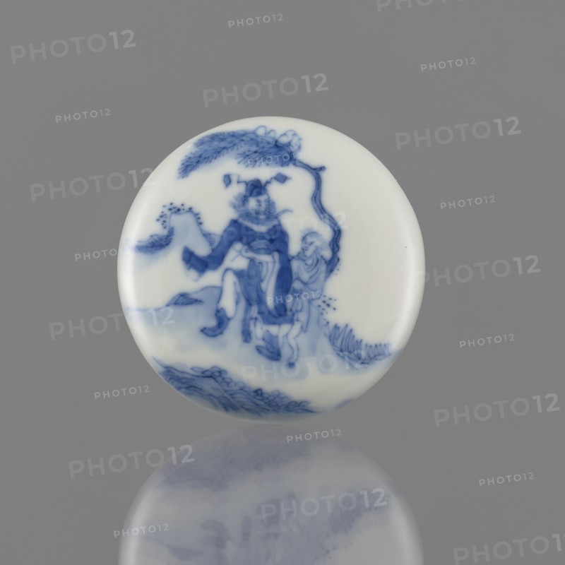 Seal ink box, depiction of Kui Xing iconography, ceramics, Chinese blue and white porcelain, writing implement, Qing dynasty 1644–1911, 19th century, China; vessel; height 2 cm, diameter 6.2 cm., A round porcelain disk with a glossy white surface decorated in blue, showing a painted scene of a figure wearing flowing garments beneath a curved tree branch, with another smaller figure nearby, stylized rocks and foliage at the base, and a soft reflection of the disk visible on the gray background; colors visible include white, blue, and gray.