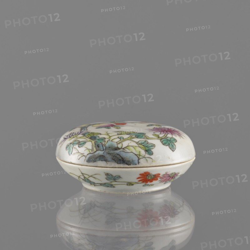 Lidded box, Chinese, colored porcelain decorated with pomegranates, chrysanthemums and peonies, floral ornaments; mid-19th century Qing dynasty, 1644–1911; ceramic vessel for writing instruments; height 3.2 cm, diameter 7.9 cm., A small oval porcelain box with a domed lid sits against a plain gray background, its surface predominantly white with painted botanical motifs in green, blue, purple, red, pink, and orange, showing leaves, flowers, and a bluish rock-like shape; the box has a thin gold-colored rim where lid meets base and rests on a reflective gray surface that creates a soft mirrored image of the painted floral decoration.