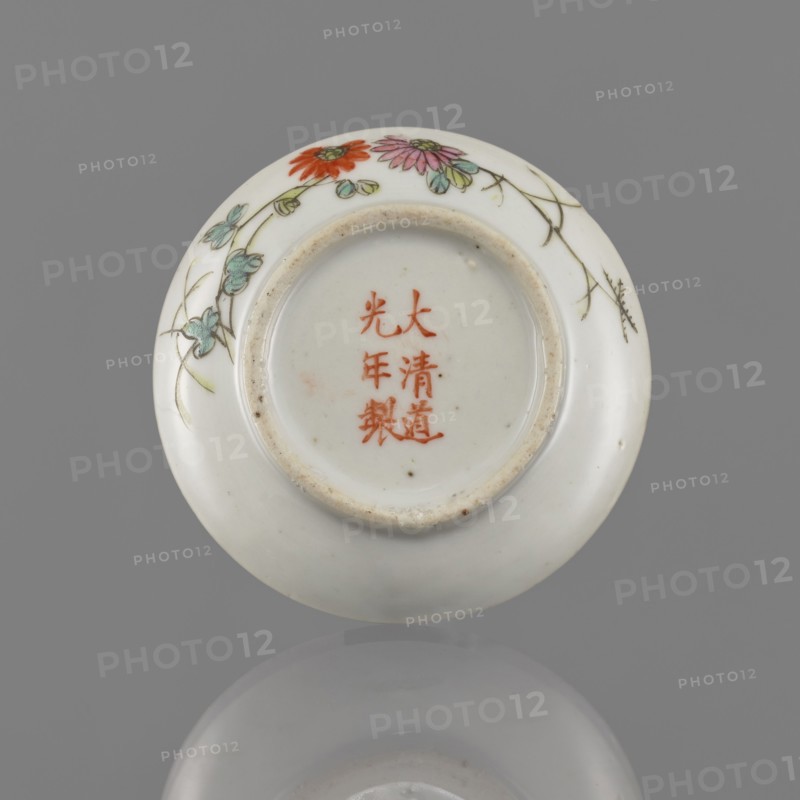 Lidded box, Chinese, colored porcelain decorated with pomegranates, chrysanthemums and peonies—floral ornaments; ceramic vessel used for writing instruments; Qing dynasty mid-19th century, 1644–1911; height 3.2 cm, diameter 7.9 cm., A round white ceramic object with a recessed base is shown flat against a gray background, decorated near the rim with painted flowers and trailing stems in colors including red, pink, green, turquoise, and pale yellow, with fine brown outline lines; the central recessed area shows a few small brown specks and the rim of the recess has a slightly rough, beige-tinged edge; the surface reflects softly onto the gray background creating a muted mirror image.