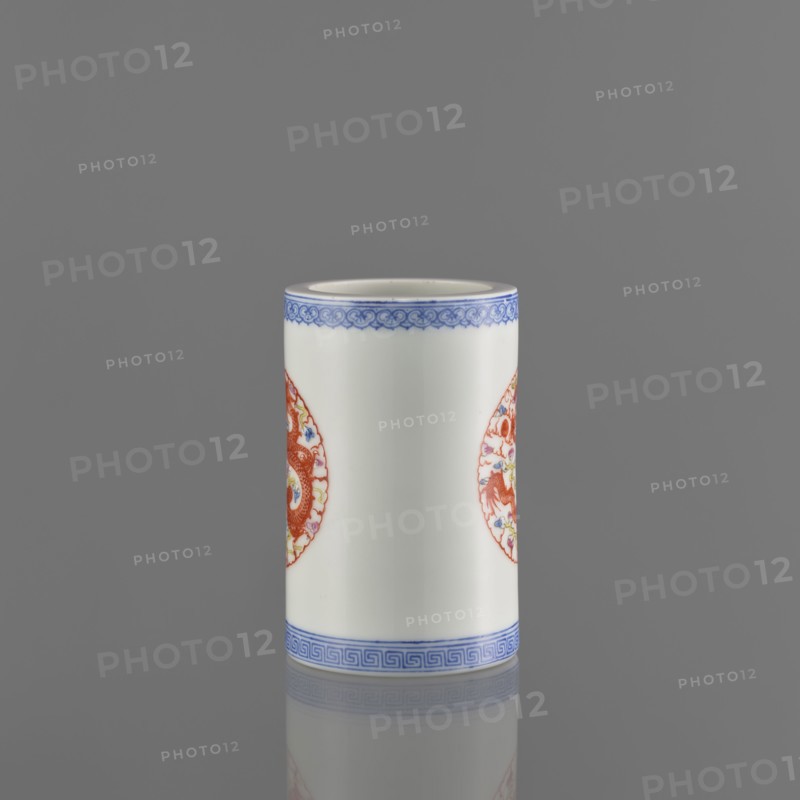 Brush vessel, Chinese, 19th century, ceramics, decorated with dragons and meander ornament, function: vessel brush holder, height 11.4 cm, diameter 7.1 cm., A cylindrical white ceramic vessel with decorative blue bands near the top and bottom edges and a pair of circular red ornamental medallions on its sides, shown standing on a reflective gray surface against a plain gray background. The visible colors include white, blue, red, and gray.