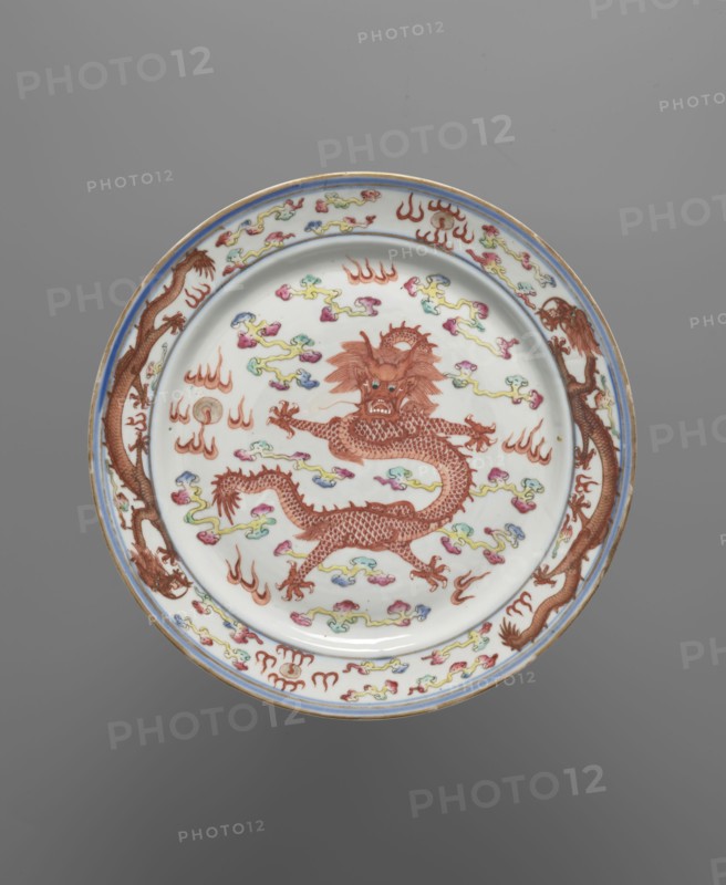 Plate, Chinese, Qing dynasty Guangxu, 1875–1908, late 19th century. Colored porcelain vessel decorated with dragons, clouds, and flames. Height 2.6 cm; diameter 25.8 cm., A round ceramic plate with a white background featuring a central red dragon with detailed scales and an expressive face, surrounded by a few cloud-like motifs in colors including yellow, green, blue, and pink; the rim has repeating red dragon figures and similar multicolored cloud elements, with a thin blue band near the edge and a narrow gold outer edge; the overall visible colors include white, red, blue, yellow, green, pink, and gold, set against a gray background.