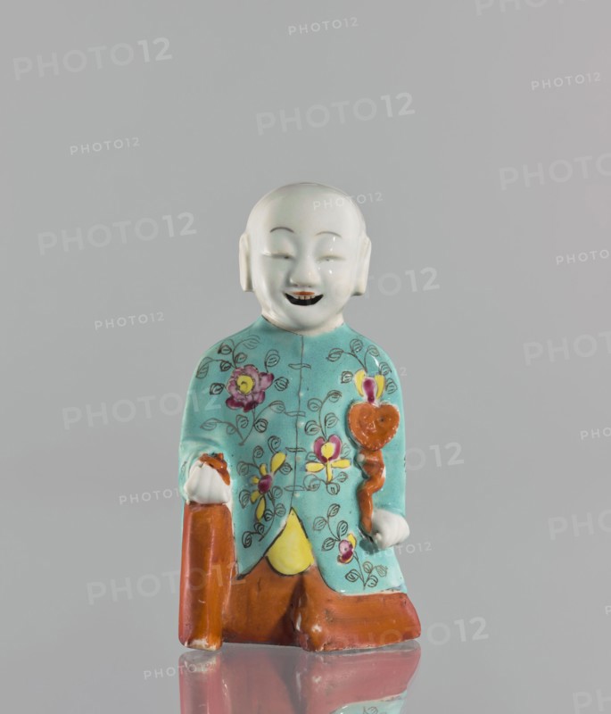Boy with ruyi — incense burner; maker, Chinese; colored porcelain; ruyi scepter of prosperity iconography; incense burner depicting a boy/child; 19th century, China; sculpture; height 15 cm., A small figurine of a smiling person with a smooth white head and facial features, wearing a turquoise garment decorated with floral patterns in pink, yellow, and green, holding a brown staff and a rounded brown object, with brown lower garments and a yellow triangular detail at the front, all set against a plain light gray background and reflected faintly on the surface beneath.