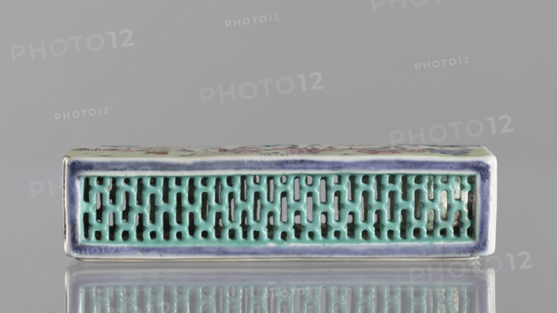 Cricket Cage, Chinese, Qing 1644–1911, colored porcelain vessel depicting animals crickets engaged in fighting/sports; 1st half of the 19th century; height 17.9 cm, width 4.6 cm., A rectangular ceramic object with a glossy finish sits horizontally against a neutral gray background, showing a front face with a patterned openwork grille in turquoise surrounded by a narrow border glazed in deep blue and white; the top edge visible has muted pastel colors and floral-like painted motifs, and the piece rests on a reflective surface that mirrors its colors and shapes.