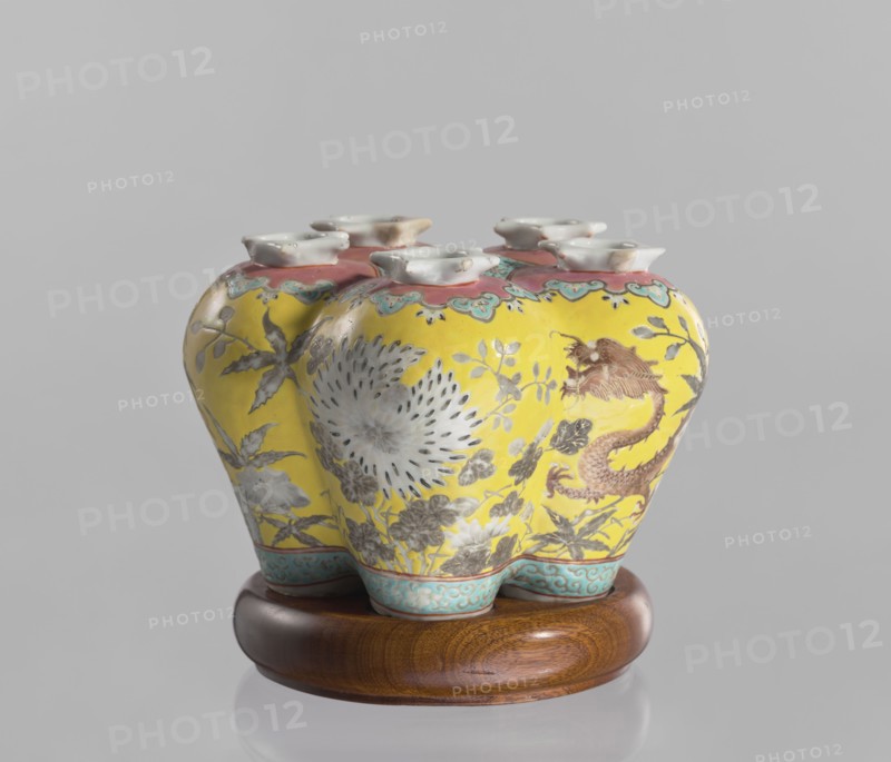 Vase in the shape of five connected vases on a base, Chinese, colored porcelain with dragons and floral decorations, ceramics, 18th/19th century China, vessel, height 15.8 cm, The image shows a ceramic vase with a bulbous, multi-lobed shape resting on a round wooden stand; the vase surface is predominantly bright yellow with painted floral and foliage motifs in muted gray and white, and a stylized dragon or serpentine creature in brownish-red visible on one side; the upper rim area has a band of coral pink with turquoise-green scalloped accents, and the feet or base edges of the vase feature a turquoise-blue patterned border; the wooden stand is warm brown with visible grain, and the background is a plain light gray.