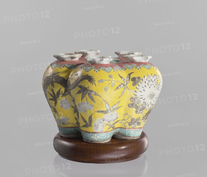 Vase in the shape of five connected vases on a base, Chinese, colored porcelain, dragons, floral decorations, ceramics, 18th/19th century, China, vessel, height 15.8 cm, An ornate ceramic vase with a bulbous, lobed body sits on a wooden stand; the vase has a bright yellow background with decorative floral and foliage motifs painted in shades of gray and white, and a large circular floral burst rendered in white and gray on one side. The top rim area is banded with a pinkish color accented by turquoise-blue scalloped patterns, and the base edge features a turquoise-blue band with a repeating decorative motif. The vase has a group of small, flared white openings at the top. The wooden stand is a rich brown with a smooth finish. Visible colors include yellow, gray, white, pink, turquoise-blue, brown, and a hint of black in the decorative detailing.