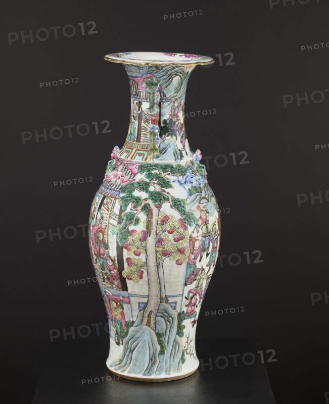 Vase, Chinese, colored porcelain, 19th century, decorated with battle scenes, figural scenes, court scenes, plant ornaments and floral decorations; vessel, height 63.2 cm, diameter 21.2 cm., A tall porcelain vase with a flared rim sits against a dark background, decorated with intricate painted scenes and raised sculptural elements; the vase shows a palette of white, green, pink, blue, gold, and brown, with areas of pale yellow and red. The body features painted trees with green foliage and pink blossoms, stylized gray-blue rocks at the base, and several groups of human figures rendered in pink, green, and brown clothing interacting around architectural elements. The neck and shoulder areas include painted buildings and terraces in green, pink, and gold tones, with small sculpted decorative motifs in pink and blue protruding from the surface. The flared rim has a gilded edge and painted floral accents in pink and green on a white background. The overall surface is glossy, and the vase ...