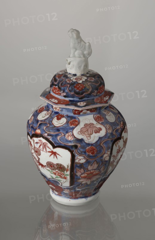 Vase with cover, Japanese, Edo period 18th century, imari porcelain decorated with flowers, tree, bamboos, peonies, and karashishi; vessel, height 25.5 cm, diameter 16 cm., A decorated ceramic jar with a lid topped by a small sculpted figure sits against a neutral gray background. The jar is predominantly blue with areas of white and red-orange, and it shows floral and cloud-like motifs in a painted pattern. Several white panels framed in dark outlines contain additional floral scenes with red-orange and green accents. The lid echoes the blue and red-orange floral design and has a white sculpted figure on top. The jar rests on a white foot and casts a faint reflection on the glossy surface beneath it.