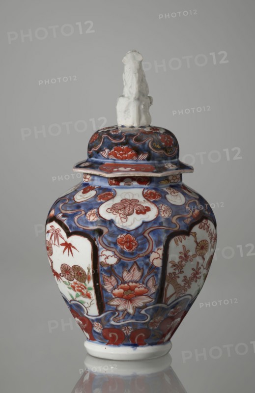 Vase with cover, Japanese, Edo period 1603–1868, 18th century; porcelain imari vessel decorated with flowers, tree, bamboos, peonies, and karashishi; height 25.5 cm, diameter 16 cm., A ceramic lidded vase with a rounded body sits against a plain light gray background; the lid is topped by a small sculpted figure in white, and the vase surface features ornate painted decoration in deep blue, red, white, and touches of green and brown, with floral motifs, cloud-like cartouches, and flowing scroll patterns covering the body and lid.