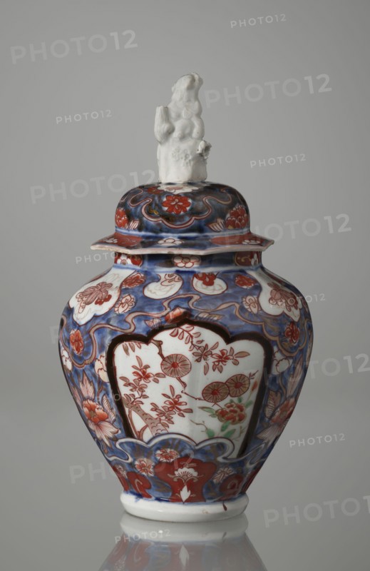 Vase with cover, Japanese, Edo period 18th century, imari ceramics decorated with flowers, tree, bamboos, peonies and karashishi lion-dog iconography; vessel, height 25.5 cm, diameter 16 cm., A lidded porcelain jar with a sculpted finial sits against a plain gray background; the finial is white and depicts a small figure with a textured surface. The jar body is bulbous with a flared rim on the lid and is decorated with an overall palette of blue, red, white, and touches of green; the dominant background decorations are blue with red floral motifs and white cartouche-shaped panels that contain red floral designs and small green accents. The surface shows swirling blue patterns, red blossoms, and stylized leaves, with a band of red and blue ornamental motifs near the base and lid, and the jar rests on a white foot that reflects faintly on the surface below.