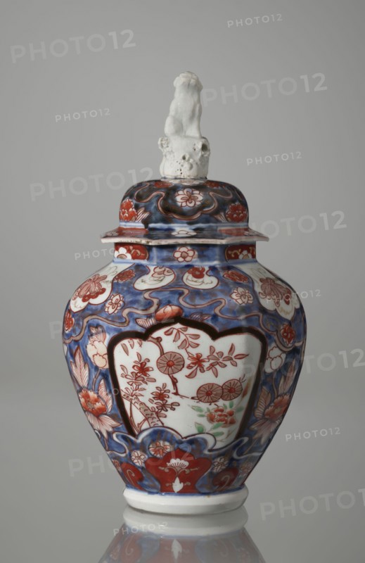 Vase with cover, Japanese, Edo period 18th century, imari ceramics decorated with flowers, tree, bamboos, peonies, and karashishi; vessel, height 25.5 cm, diameter 16 cm., A lidded ceramic vase with a glossy finish sits against a neutral gray background, featuring a predominantly blue and red painted surface with white areas left unpainted; the body is bulbous with a narrow base and a flared rim supporting a domed lid topped by a small white sculpted figure. The painted decoration includes flowing blue waves and cloudlike shapes, red floral motifs and stylized petals, and white panels on the body containing red botanical designs with touches of green. The lid echoes the blue and red floral patterning, and the small sculpted finial is textured and off-white. The base and rim show unpainted white ceramic, and the overall color palette visible is blue, red, white, green, and off-white.