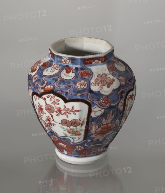 Vase with cover, Japanese, Edo period 18th century, imari porcelain vessel decorated with flowers, tree, bamboos, peonies and karashishi; height 25.5 cm, diameter 16 cm., A rounded ceramic vase with an open top sits on a reflective surface, decorated with flowing blue background patterns and panels outlined in dark tones. The vase shows floral and leafy motifs painted in red and white within scalloped and cloud-like shapes, and there are accents of green among some floral elements. The rim and base are primarily white with slight discoloration at the inner rim, and the overall visible colors include blue, red, white, green, and dark brown.