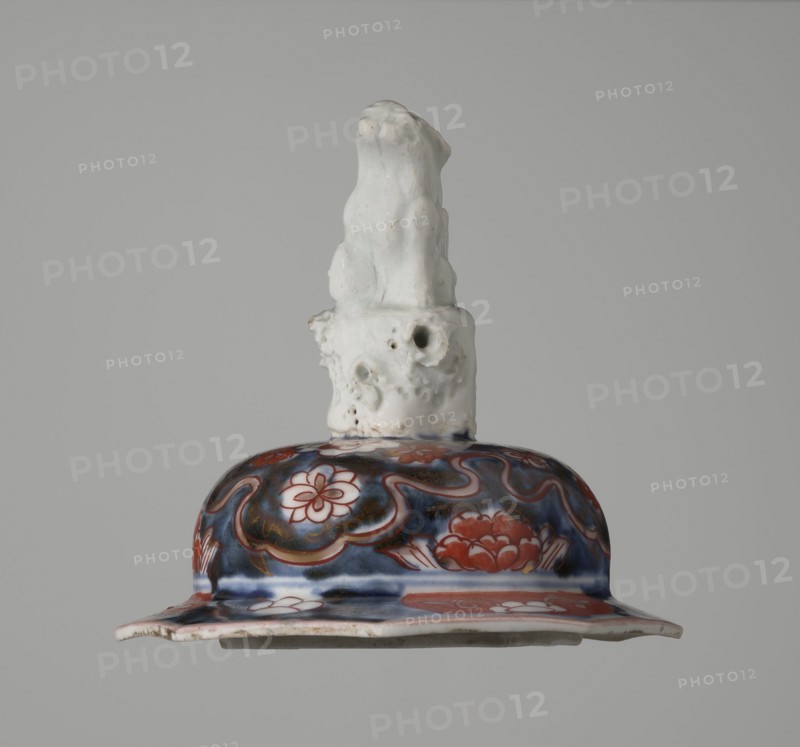 Vase with cover, Japanese, Edo period 18th century, imari ceramics depicting flowers, tree, bamboos, peonies, and karashishi; vessel; height 25.5 cm, diameter 16 cm., A ceramic object with a glossy white sculpted top resembling an irregular organic form sits above a rounded decorative section; the upper portion is matte white with some small pits and rough texture, while the lower rounded area displays a glossy patterned surface in deep blue and red with floral motifs and curving lines, and a flared rim showing white and red elements and some worn edges.