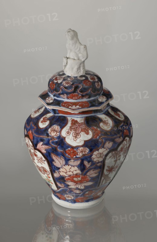 Vase with cover, Japanese, Edo period 18th century, imari ceramics decorated with flowers, tree, bamboos, peonies and karashishi; vessel, height 25.5 cm, diameter 16 cm., A ceramic lidded vase with a glossy surface sits against a plain light gray background, the body and lid covered in an intricate floral pattern in deep blue and reddish-orange with areas of white showing through; the lid is topped by a small white sculpted figure. The vase has a rounded shoulder that tapers toward the base, and the decoration includes stylized flowers and leaves, curving outlines, and panel-like white medallions framed by the blue and reddish-orange motifs. The overall color palette visible is deep blue, reddish-orange, white, and small touches of pale green, with subtle reflections of the vase visible on the glossy surface beneath it.