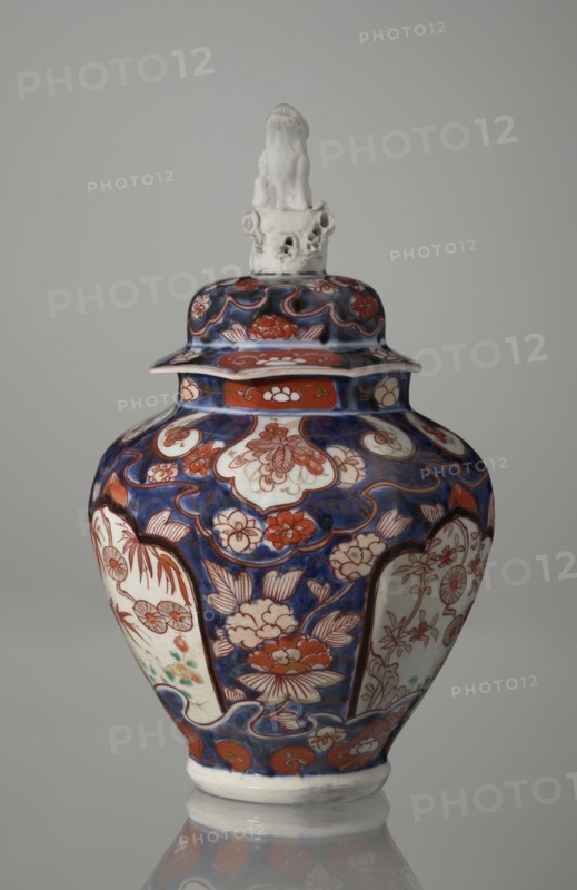 Vase with cover, Japanese, Edo period 18th century, imari ceramics decorated with flowers, tree, bamboos, peonies, and karashishi; vessel, height 25.5 cm, diameter 16 cm., A lidded ceramic vase with a rounded body and narrow base sits against a plain light gray background, decorated in a palette of deep blue, red, white, and touches of green; the surface shows a repeating floral motif with stylized blossoms and leaf patterns outlined in red against blue fields and framed white panels, while the lid features similar blue and red ornamentation topped by a sculpted white finial resembling an abstract organic form.