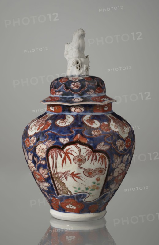 Vase with cover, Japanese, Edo period 18th century, imari ceramics depicting flowers, bamboo, peonies, and karashishi; vessel, height 25.5 cm, diameter 16 cm., A ceramic lidded jar with a glossy surface, featuring a deep blue background with extensive red and white floral and cloud-like motifs, and accents of green and brown in the painted panels; the jar has a bulbous body tapering to a narrow base with a white foot, a flaring rim under the lid, and a lid topped by an irregular white sculpted finial that has a rough, textured appearance; the painted decoration includes framed panels on the body with stylized botanical scenes in red and brown on a white ground, surrounded by red and white flowers and swirling blue patterns, and the overall color palette visible is blue, red, white, green, brown, and a hint of black in the outlining.