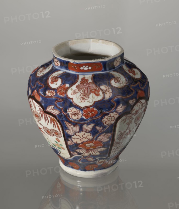 Vase with cover, Japanese, Edo period 18th century, imari porcelain vessel decorated with flowers, tree, bamboos, peonies, and karashishi; height 25.5 cm, diameter 16 cm., A rounded ceramic vase with a wide mouth sits against a neutral gray background, decorated with a deep blue ground and a pattern of floral shapes and curved panels in shades of red, white, and a touch of green. The vase shows several stylized flowers and leaves in red and white set within blue areas, with white panels bordered by red outlines that contain more floral designs. The rim and base are white with some red decorative elements near the top band, and the overall surface has a glossy finish that creates a soft reflection on the surface beneath it.