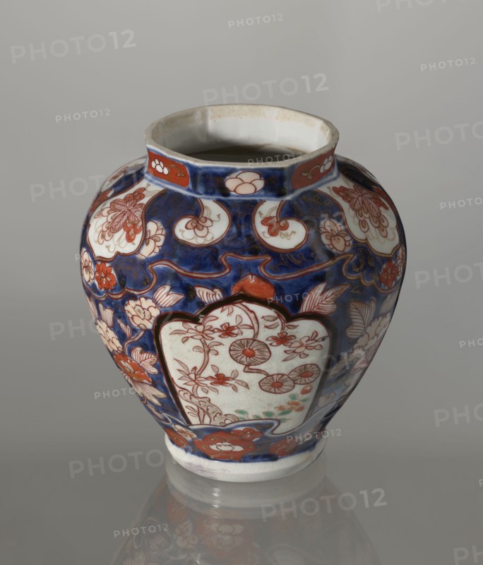 Vase with cover, Japanese, Edo period 18th century, imari ceramics, decorated with flowers, tree, bamboos, peonies, and karashishi lion-dog iconography; vessel height 25.5 cm, diameter 16 cm., An ornate ceramic vase with a rounded body and a short neck sits on a reflective surface, displaying a dominant deep blue background with decorative areas of white and red-orange, plus touches of green and pale beige; the surface is covered in floral and foliate motifs including stylized blossoms, leaves, and scrolling forms outlined in dark lines, and several white cartouche-like panels containing delicate red-orange floral drawings; the neck has a band of red-orange panels with small white floral shapes, and the rim and interior show off-white and beige tones.