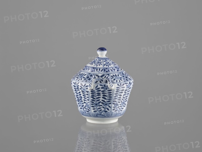Sugar bowl with lid, ceramics, sugar bowl, Chinese art, Chinese culture, lid, 20th century, China, vessel, height 12 cm, A small lidded ceramic container sits centered on a reflective surface against a plain gray background; the container is white with detailed blue floral and geometric patterns covering its body and lid, and it has a rounded finial on top with blue accents and a small curved handle on the side, with the blue-and-white motif mirrored faintly in the reflection below.