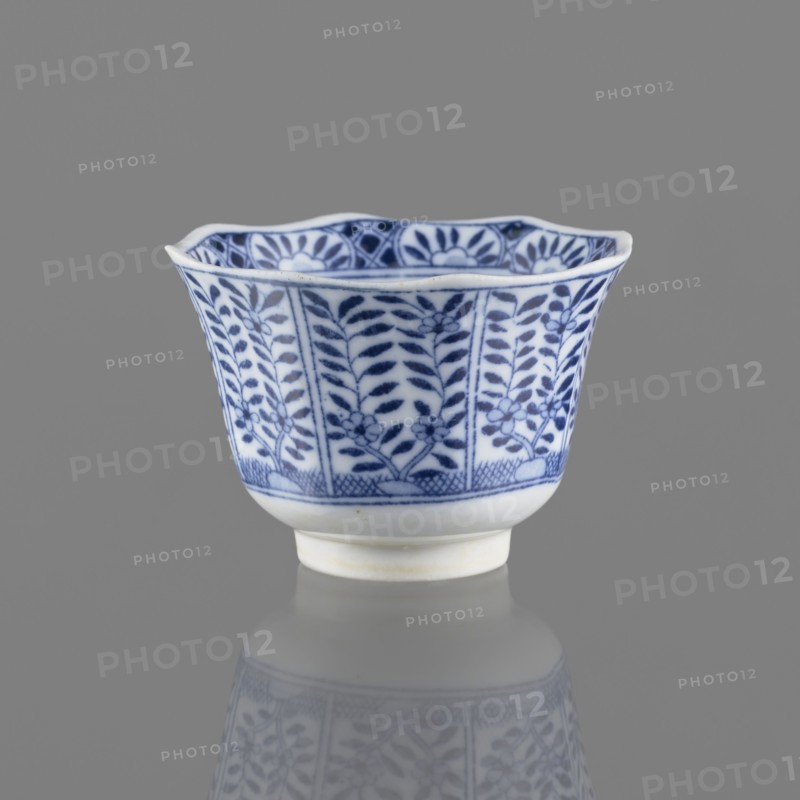 Bowl, Chinese, ceramics, 20th century, China, vessel; height 5.3 cm, diameter 7.8 cm, A small porcelain cup with a scalloped rim sits against a neutral gray background, decorated with a repeating blue floral and leafy pattern on a white ground, the blue designs appearing darker in some areas and lighter in others; the cup has a slightly flared shape narrowing to a short foot, and a soft reflection of the cup is visible on the glossy surface below.