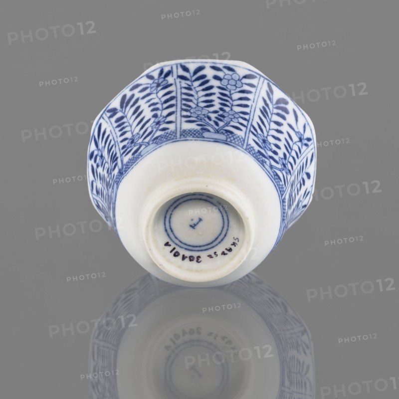 Bowl, Chinese, 20th century, ceramics vessel, height 5.3 cm, diameter 7.8 cm, A small ceramic cup is shown against a gray background, positioned rim-up so the base faces the viewer; the cup is white with a blue floral and leaf pattern around its exterior, featuring vertical panels of stylized leaves and flowers in dark blue on a white ground, and a narrow band of crosshatching near the rim. The base is recessed and plain white with a few concentric blue rings visible inside the foot, and the cup casts a soft reflection on the glossy surface beneath it. Colors visible include white, dark blue, and gray.