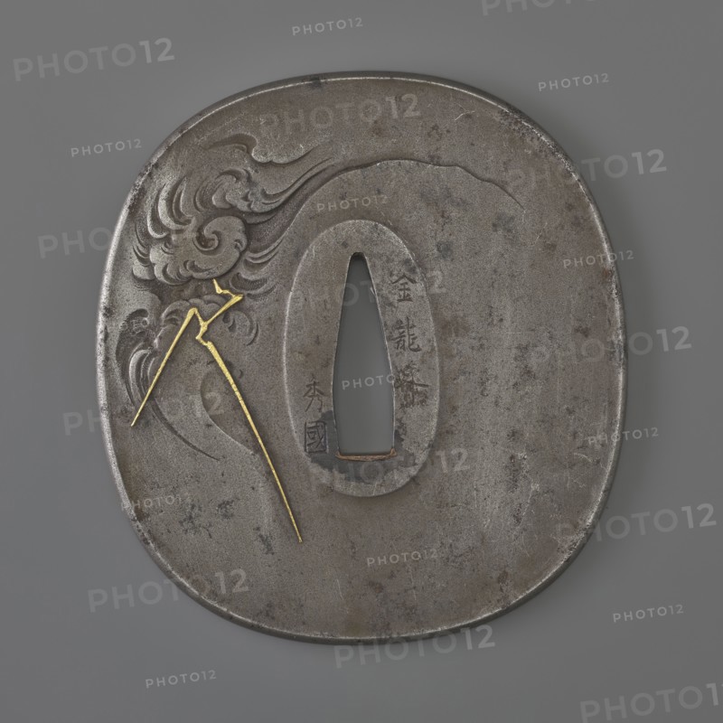 Crossguard tsuba by Kawarabayashi Hidekuni 1825–1891. Rectangular tsuba with rounded corners nadegaku gata and a slightly raised, rounded edge of the uchi nkaeshi mimi type. Decorated with a dragon with a pearl among clouds with lightning, executed in painterly engraving imitating ink painting. Details inlaid with gold and very fine agates. Signed. Hidekuni was a student of Hideoki, a master of the Ôzuki school; he died in 1891. 2nd half of the 19th century Edo/Meiji periods, Japan. Tsuba; height 7.9 cm, width 7.3 cm, depth 0.4 cm., A round metal object with an oval hole near its center sits against a gray background; the metal surface is mostly dark gray with areas of lighter gray and brownish spots, and a carved flowing cloud-like pattern appears on one side. A thin bright gold line in a few segments is inlaid across the carved area, forming angular shapes that contrast with the gray metal. The object shows subtle surface scratches and varied patina, and its edges are slightly irregu...