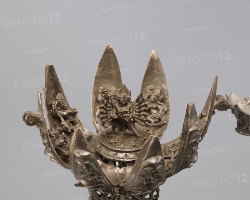 Lotus flower with Buddhist deities, 19th century, Nepal, bronze cult object in the form of a lotus mandala depicting Buddhist deities bodhisattva iconography, lotus iconography, pairs of lovers, enlightenment, height 20.3 cm, width 8.0 cm, A metallic sculptural object shaped like an open lotus with a central figure standing on a round pedestal, surrounded by several petal-like panels each bearing smaller relief figures; the metal has a bronze or brass appearance with brown and gold tones and areas of darker patina; the petals curve upward and outward, showing carved faces and figures in low relief; the central figure has multiple arms arranged in a radial pattern and a detailed headdress; the background is a plain light gray.