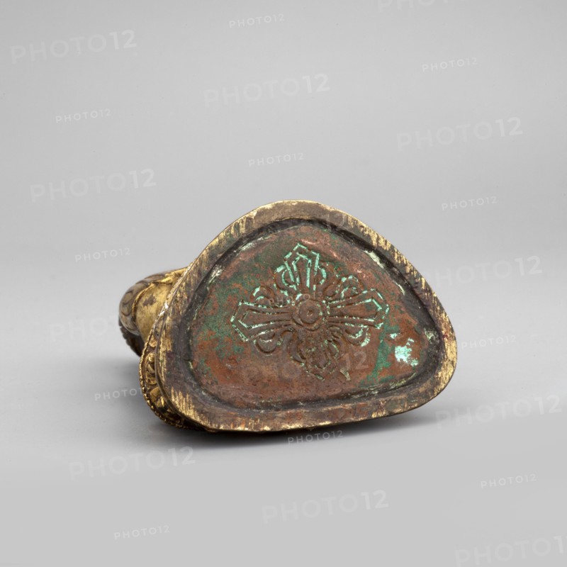 Shakyamuni Buddha, Buddha iconography, Himalayan/Tibetan Buddhist art, Buddha ca. 560–480 BCE, ushnisha, 19th century, Tibet, sculpture figurine; height 6.7 cm, width 3.3 cm, depth 4.1 cm., A small decorative object rests on a neutral gray surface against a light gray background, shown from a low frontal angle revealing its irregular triangular base with a raised floral-like pattern in the center; the object shows a worn metallic finish with prominent gold tones on its rim and sides, areas of brownish copper coloration across the base, and patches of greenish patina on and around the carved design, with subtle darker and lighter variations in the surrounding metal.