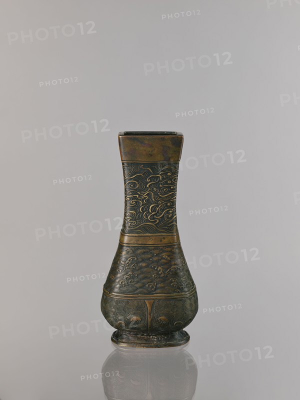 Hu-type vase, relief decoration, hu vessel, Chinese art, Chinese culture, meander ornament, ceremonial vessels, clouds, sea waves, ritual vessels, Xuande 1426-1435, Ming 1368-1644, Qing 1644-1911, backdated signatures, quadrilateral with rounded lower edges, with an inscription, 17th century, China, vessel, height 24.0 cm, width 12.0 cm, A single ornate vase stands against a plain light gray background, showing a tall narrow neck that widens into a rounded base with a small foot; the vase has a metallic appearance with dark bronze and muted gold tones, and its surface is covered in carved swirling patterns and wave-like motifs that repeat across the neck and body, with bands separating the sections and a subtle reflective sheen that creates darker and lighter areas on the metal.