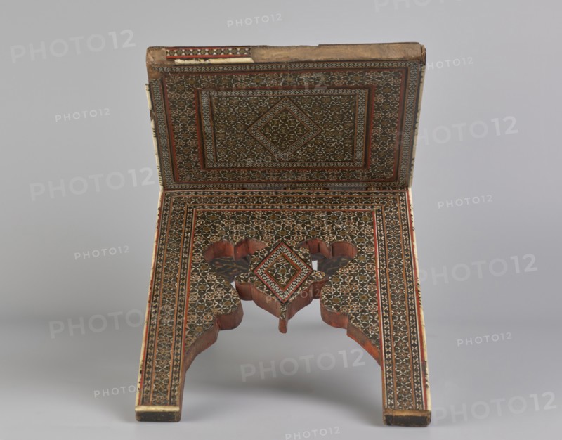 Quran lectern rahla, Islamic art, Persian art, Qajar dynasty 1785–1925, geometric ornament, stands, rahla, Quran, Persian culture, 19th century, Iran, piece of furniture; unfolded: length 21.5 cm, width 41.4 cm; folded: length 41.5 cm, width 16.5 cm., An ornate folding wooden stand with intricate geometric inlaid patterns covering its surfaces, showing a rectangular top panel hinged to an angled lower panel that splits into two carved legs; the patterning forms concentric framed designs with a central diamond motif on both panels, and the lower panel includes a decorative cutout where the legs meet; visible colors include brown from the wood, dark brown or black in the inlay, red accents, cream or beige highlights, and small touches of green, all set against a neutral light gray background.