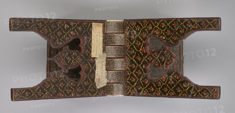 Quran lectern rahla, Islamic/Persian art, Qajar dynasty 1785–1925, geometric ornament, stand for Quran, Persian, 19th century, Iran, piece of furniture; unfolded: length 21.5 cm, width 41.4 cm; folded: length 41.5 cm, width 16.5 cm., An open ornate object with mirrored symmetrical halves connected by a central ribbed spine, each half covered in a dark background patterned with winding yellow vines and small red floral motifs, edged with thin red trim; the inner surfaces show darker inset panels with intricate geometric designs in muted browns and grays, and a few irregularly shaped cutouts revealing more patterned interior layers; a narrow vertical strip of light-colored worn material is attached near the spine with visible rough edges; the overall color palette includes black, yellow, red, brown, gray, and beige.