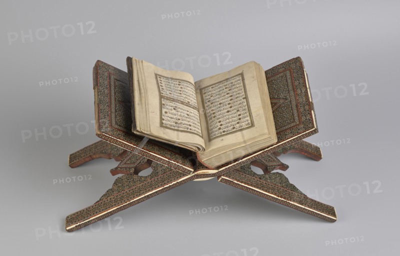 Quran lectern rahla, Islamic art, Persian art, Qajar dynasty 1785–1925, geometric ornament, stands, rahla, Quran, Persian culture, 19th century, Iran, piece of furniture; unfolded: length 21.5 cm, width 41.4 cm; folded: length 41.5 cm, width 16.5 cm., An open book with aged, off-white pages rests on an ornately carved wooden stand with an X-shaped support; the stand shows detailed geometric and floral patterns in dark brown, reddish-brown, and cream with accents of green and gold; the book's pages are cream with visible lines of script and decorative elements in muted brown and faded black; the background is a smooth light gray.
