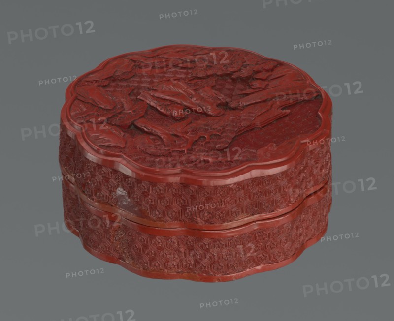Box with a lid; creator, Chinese; Qing dynasty 1644–1911; 2nd half of the 18th century. Vessel, height 11.0 cm, width 4.8 cm. Materials and/or decoration: figurative scenes of sages, scholars, and servants; landscape motifs with pine trees and rocks; rosettes; geometric ornament including kikko pattern., The image shows a round, scalloped-edge carved box in a deep red color against a muted gray background. The top surface features raised, textured carving with organic, leaf-like shapes and layered relief. The sides display a repeating hexagonal geometric pattern in low relief, with smooth raised bands encircling the box separating the top and bottom sections. The red finish varies slightly in tone, with some areas appearing darker and others slightly lighter, and there are small patches where the surface shows a different sheen. The background is a flat, even gray.