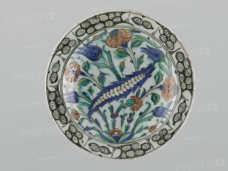 Plate, Islamic art, Turkish, Ottoman dynasty 1299–1922, floral motifs including tulips, carnations, saz, early 17th century, Iznik, Turkey; vessel; height 29.0 cm, width 5.7 cm., A round ceramic plate with a decorative floral design fills the frame, showing a central motif of stylized flowers and leaves in shades of blue, green, and orange on a white background, with a prominent curved blue element edged with a white bead-like pattern. The rim features a repeating black spiral pattern on white, and the inner field contains several tulip-like shapes, rounded blossoms, and slender stems with leaves in green and turquoise, plus small orange flower accents. The overall colors visible are white, blue, green, black, orange, and a touch of turquoise.