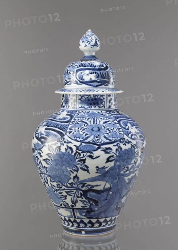 Vase with cover, unknown ceramist, Japanese, Edo period Genroku–Kyoho, ca. 1690–1730, Arita porcelain, blue-and-white; decorated with phoenixes, hares/rabbits, peonies, chrysanthemums, and lotuses; one of the four elements of a fireplace set; height 59.0 cm, diameter 33.0 cm., A lidded porcelain vase standing against a plain background, decorated in blue and white. The vessel has a bulbous body tapering to a narrower base and a matching lid topped with a pointed finial. The surface shows dense painted ornamentation including floral motifs, leafy branches, stylized panels, and a bird in flight among the foliage. The blue decoration varies in tone from deep indigo to lighter blue washes, set against a white ground. The lid and neck carry coordinated patterned bands and small decorative panels, and the base has a repeating border motif. The background color is a neutral gray.