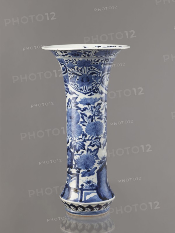 Vase, Japanese, Edo period 1603–1868, Genroku–Kyoho ca. 1690–1730. Blue-and-white porcelain from Arita, decorated with hares/rabbits, peonies, lotuses, and phoenixes kacho/kachôga motifs. One of four elements of a set; vessel height 39.0 cm, diameter 20.3 cm., A tall, flared ceramic vase with a glossy white base decorated in blue floral and foliate patterns, featuring large stylized flowers and leaves arranged vertically along its narrow body, a flared rim with decorative bands and motifs, and a slightly flared foot with a repeating decorative border; the background is a smooth, neutral gray, and the vase shows shades of deep blue and lighter blue on white with subtle reflections on its surface.