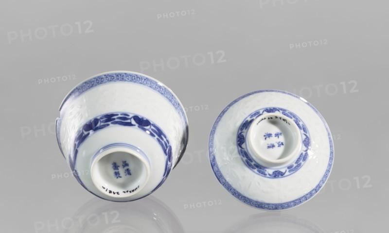 Bowl on a base with a lid, ceramic, rice porcelain, geometric ornament, rice pattern, plant ornament, Chinese art/culture, Qing dynasty 1644–1911, late 18th century, vessel, height 10 cm. Floral arrangement in a circle at the bottom. Signed., A pair of porcelain vessels shown from above and slightly tilted, each painted with blue decorative bands and motifs on a white background; one vessel has a small chip on the rim, and both show glossy surfaces and circular openings with visible recessed bases; the color palette is primarily white and blue with a neutral gray background and faint reflections beneath the objects.